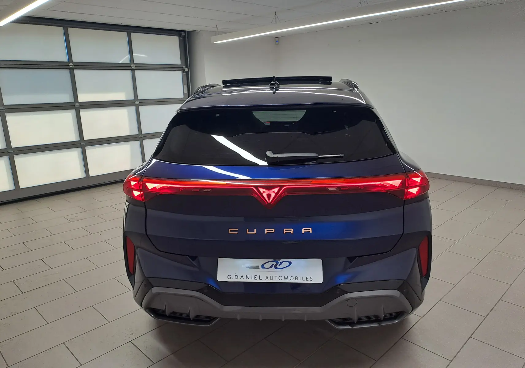 Vue arrière d'une CUPRA TERRAMAR bleu cosmos avec feux arrière allumés et toit ouvrant panoramique en intérieur.