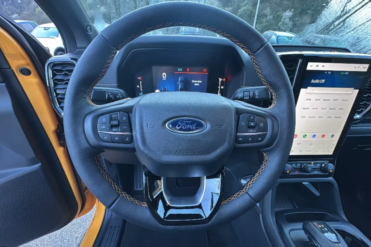 Vue intérieure centrée sur le volant cuir noir avec surpiqûres orange du Ford Ranger Cyber orange, écran tactile et tableau de bord numériques visibles.