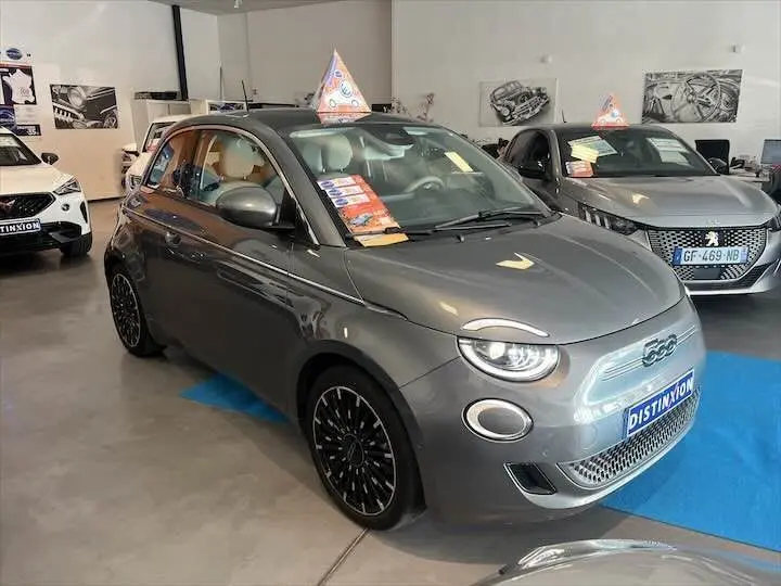 Fiat 500 électrique gris minéral métal vue 3/4 avant en showroom avec drapeau publicitaire sur le toit.