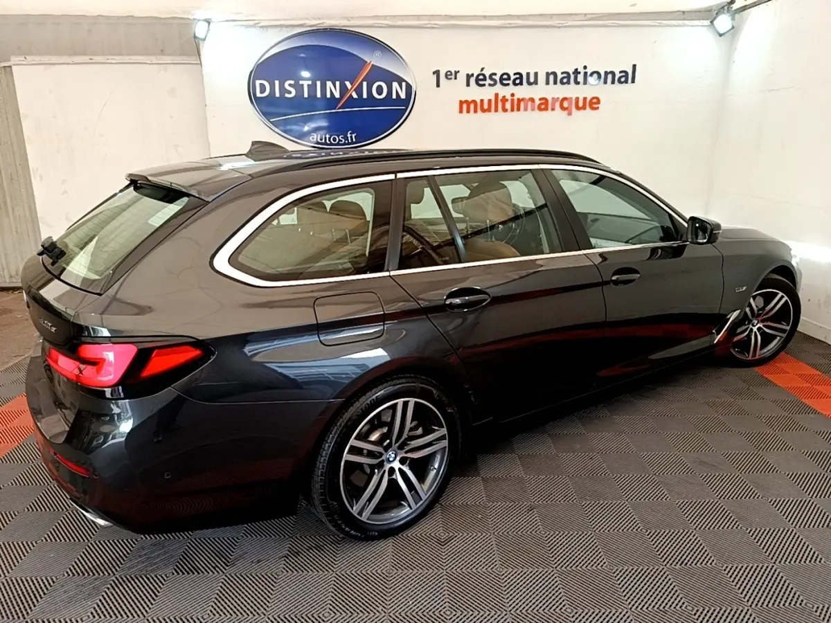 BMW Série 5 Touring 530e gris clair vue 3/4 arrière droit, avec jantes alliage et feux arrière LED allumés.