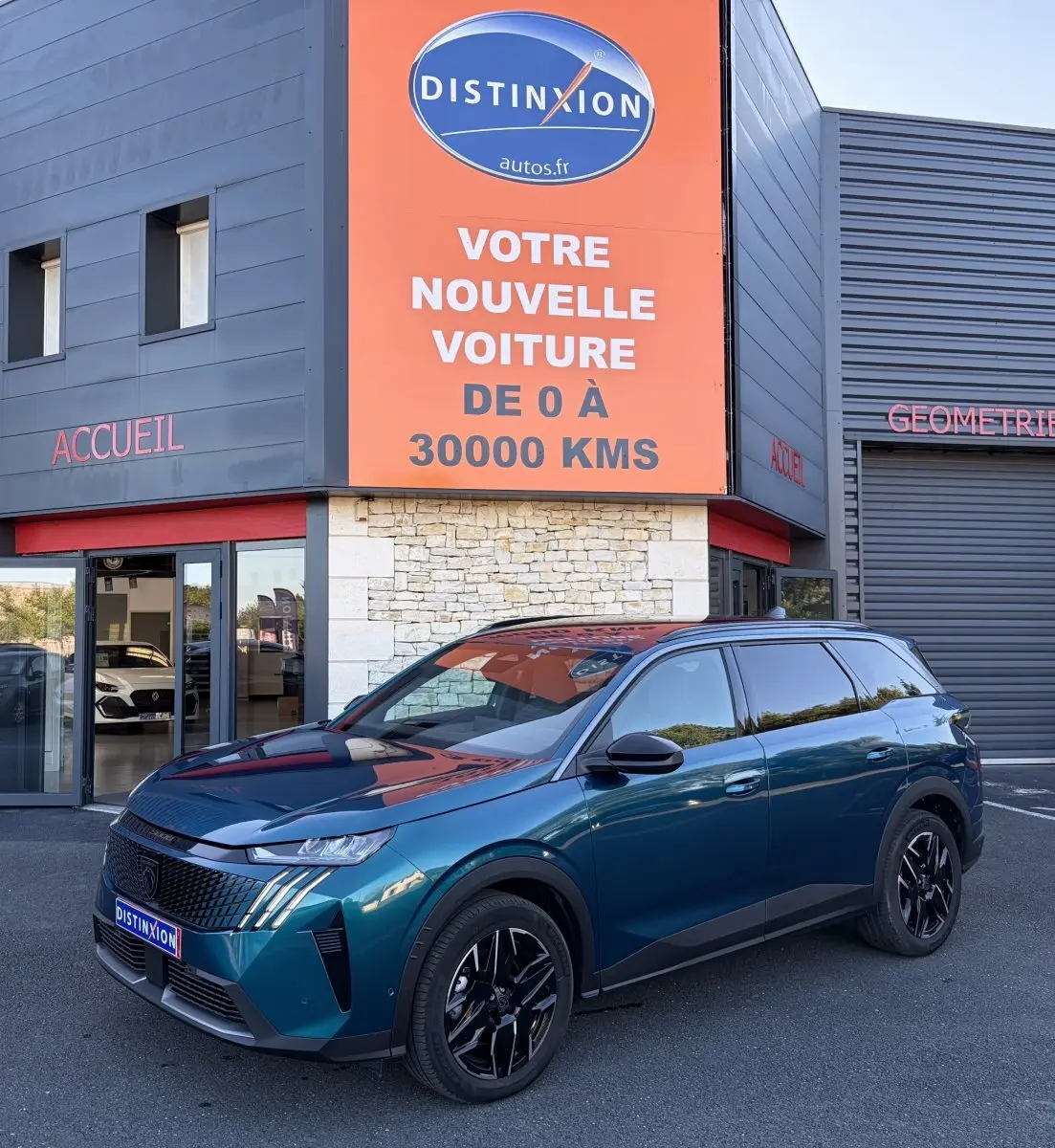 Peugeot 5008 hybride vert en 3/4 avant droit, avec jantes noires et calandre distinctive, devant un bâtiment commercial.