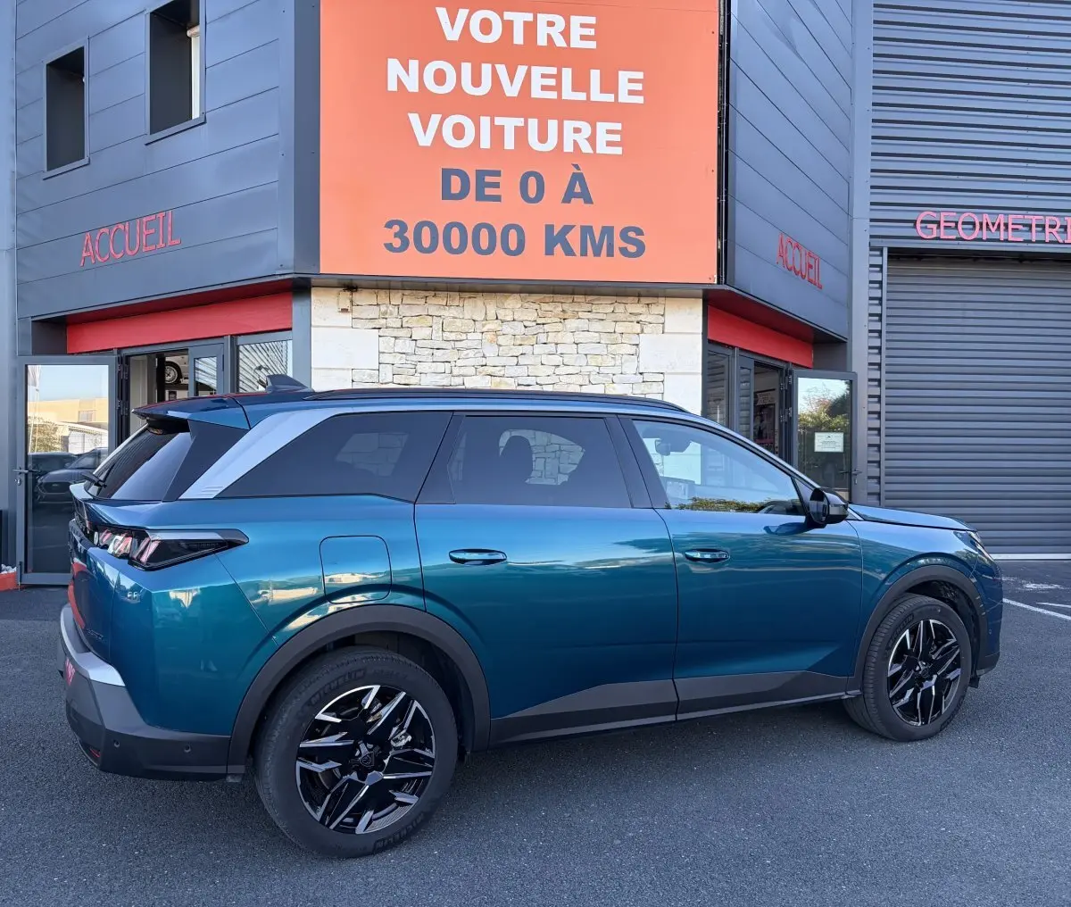 Vue côté gauche d’un Peugeot 5008 hybride vert 2025, avec jantes noires et toit noir, stationné devant un bâtiment commercial.