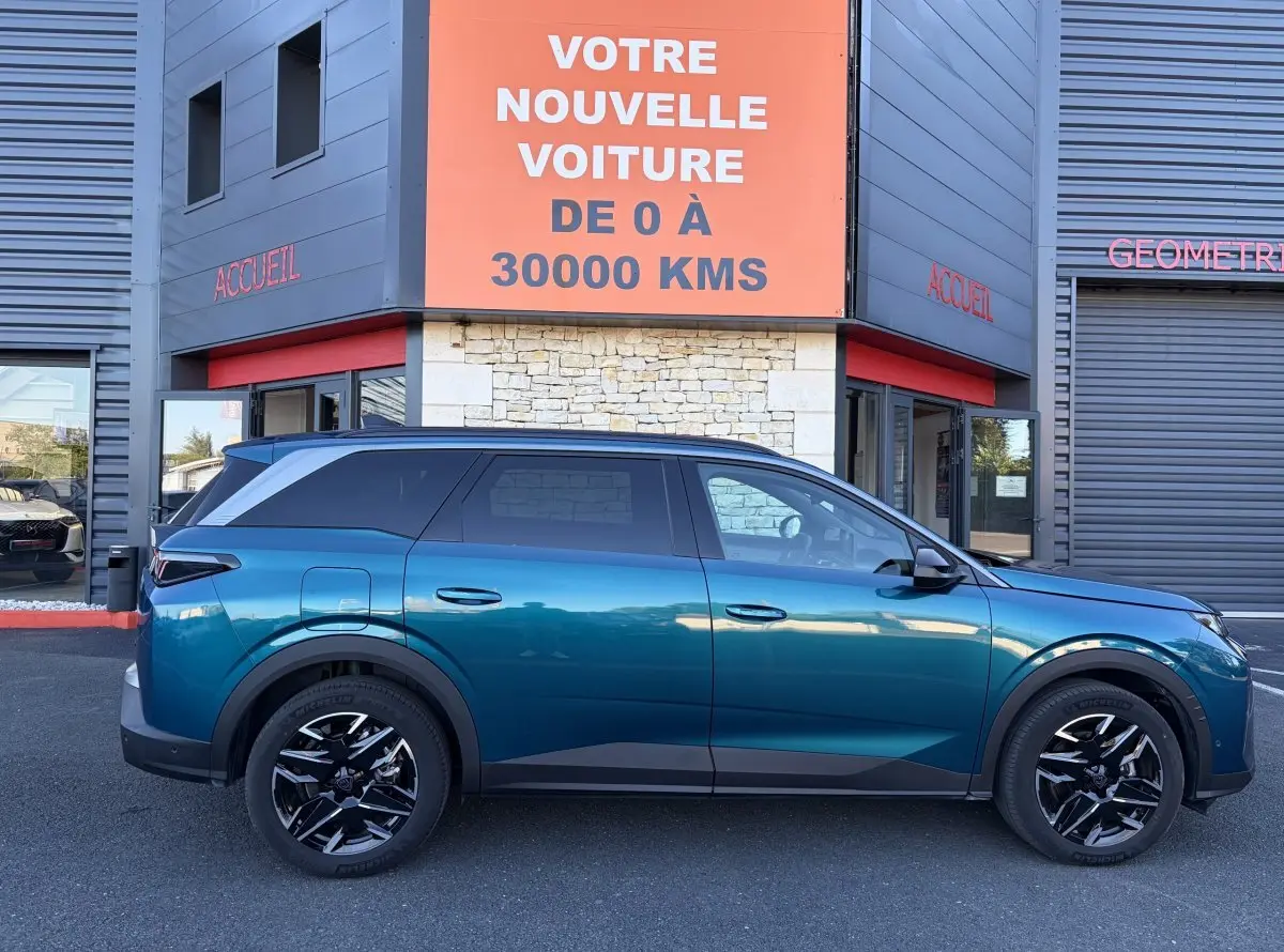 Profil côté gauche d’un Peugeot 5008 hybride vert métallisé, garé devant un bâtiment avec panneau orange.