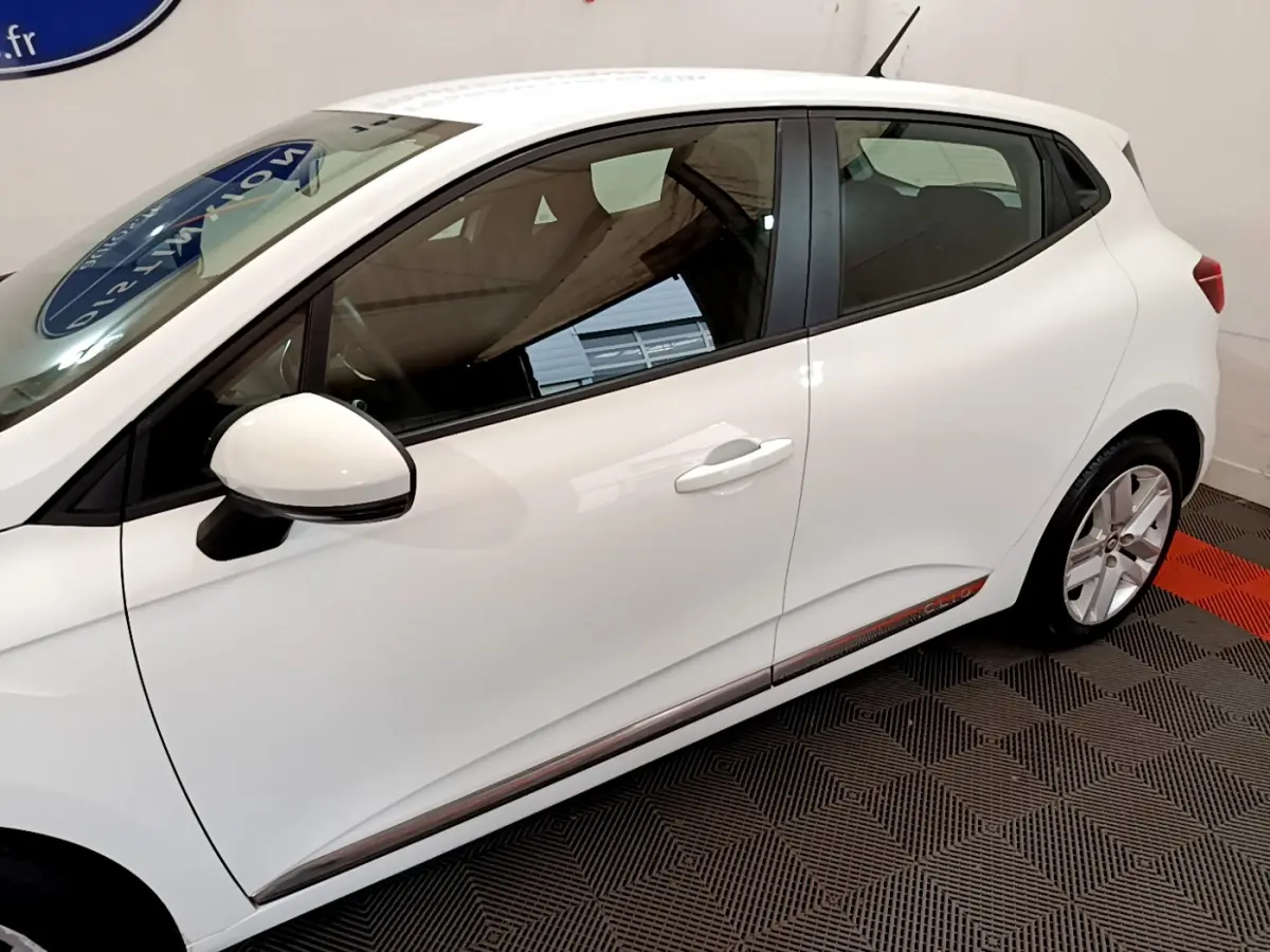 Profil côté gauche d'une Renault Clio Business TCe 100 blanche avec rétroviseur et poignées assortis, sur sol damier noir et rouge.