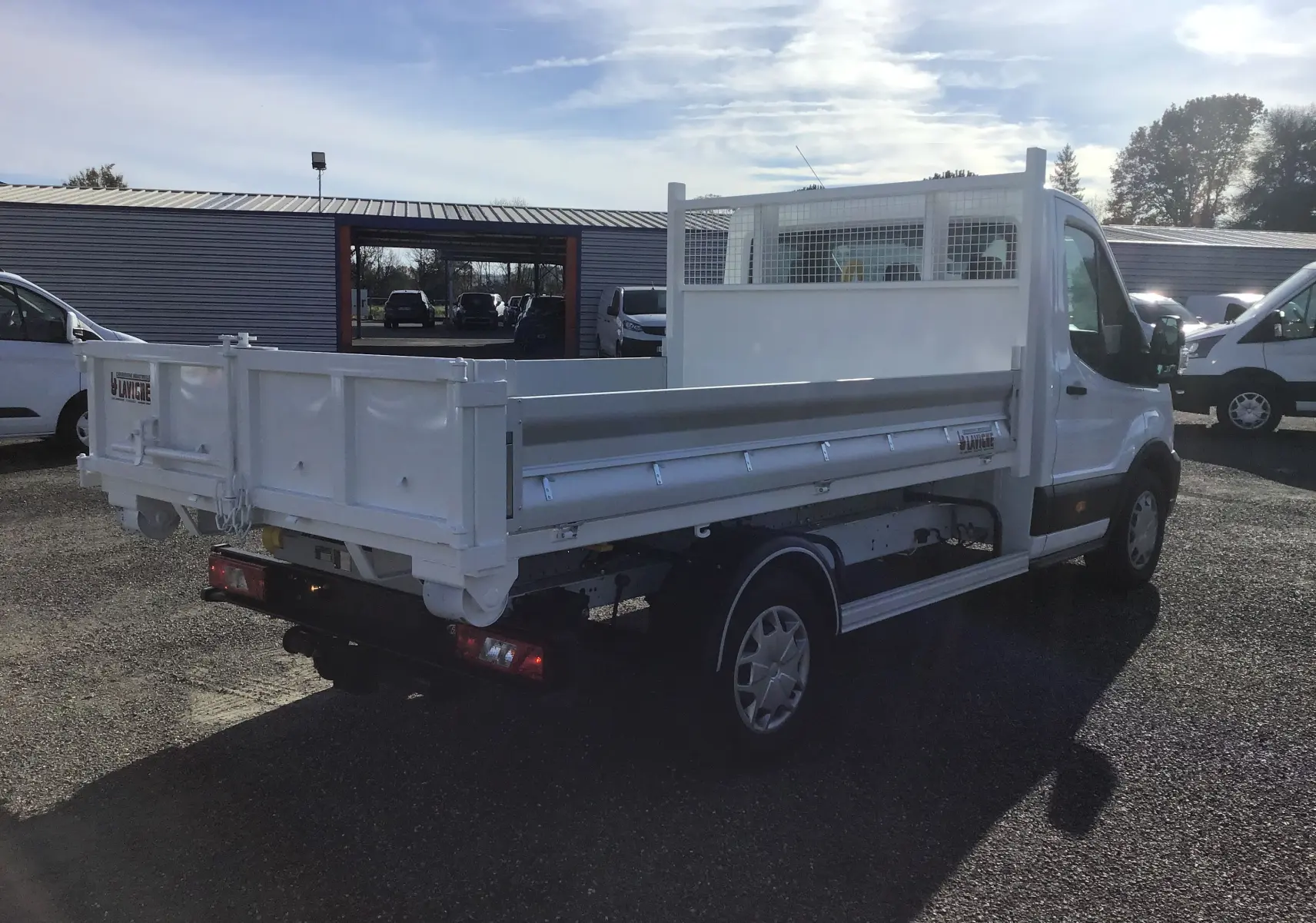 Ford Transit Chassis cabine blanc vu en 3/4 arrière droit avec benne 3m20 et bras polybenne sur parking.