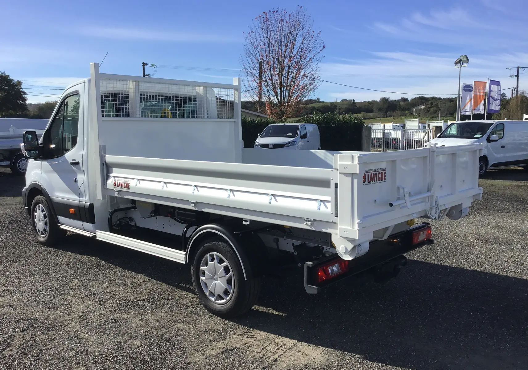 Ford Transit Chassis Cabine blanc vu en 3/4 arrière droit avec benne basculante et bras polybenne sur parking extérieur.