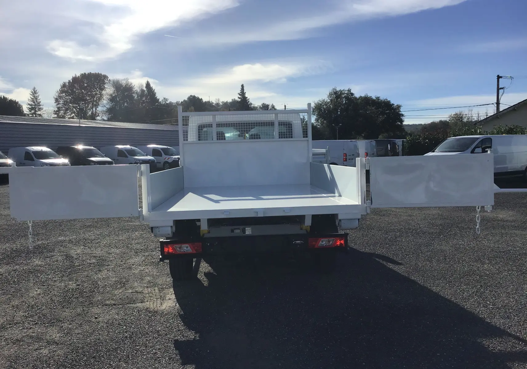 Vue arrière d'un Ford Transit Chassis Cabine blanc avec benne ouverte et portes latérales déployées sur un parking extérieur.