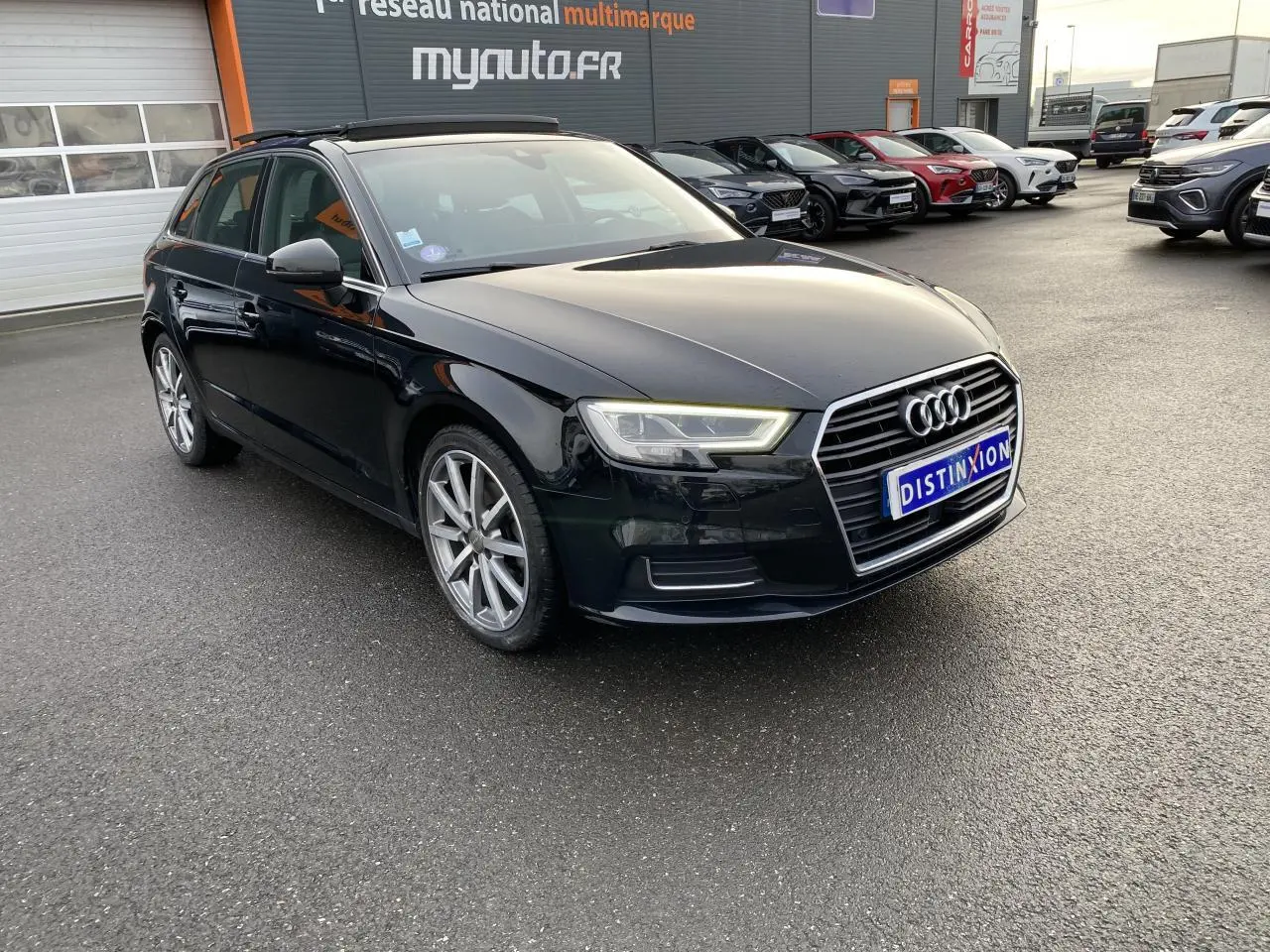 Audi A3 Sportback noir vue 3/4 avant droit, avec phares LED allumés sur parking extérieur.