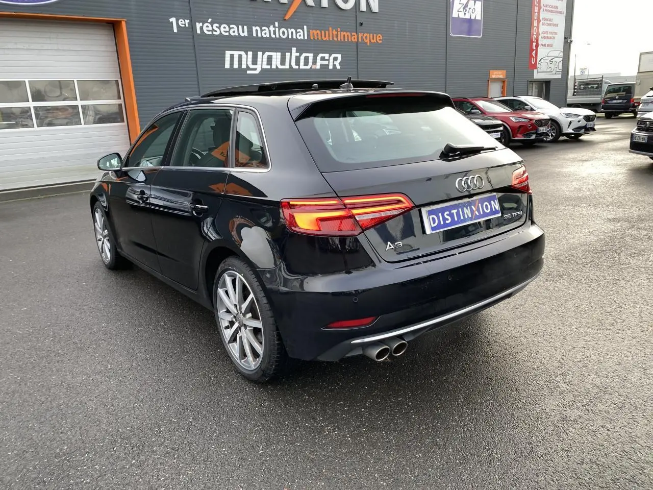 Audi A3 Sportback noire vue 3/4 arrière droit, feux arrière LED allumés et double sortie d’échappement chrome.