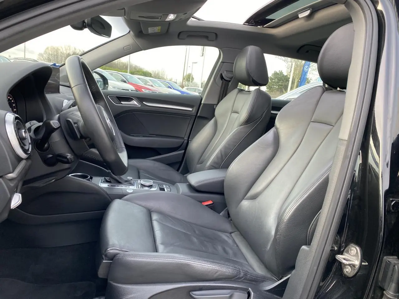 Vue intérieure côté conducteur de l'Audi A3 Sportback noire, montrant les sièges sport en cuir Milano et le toit panoramique en verre.