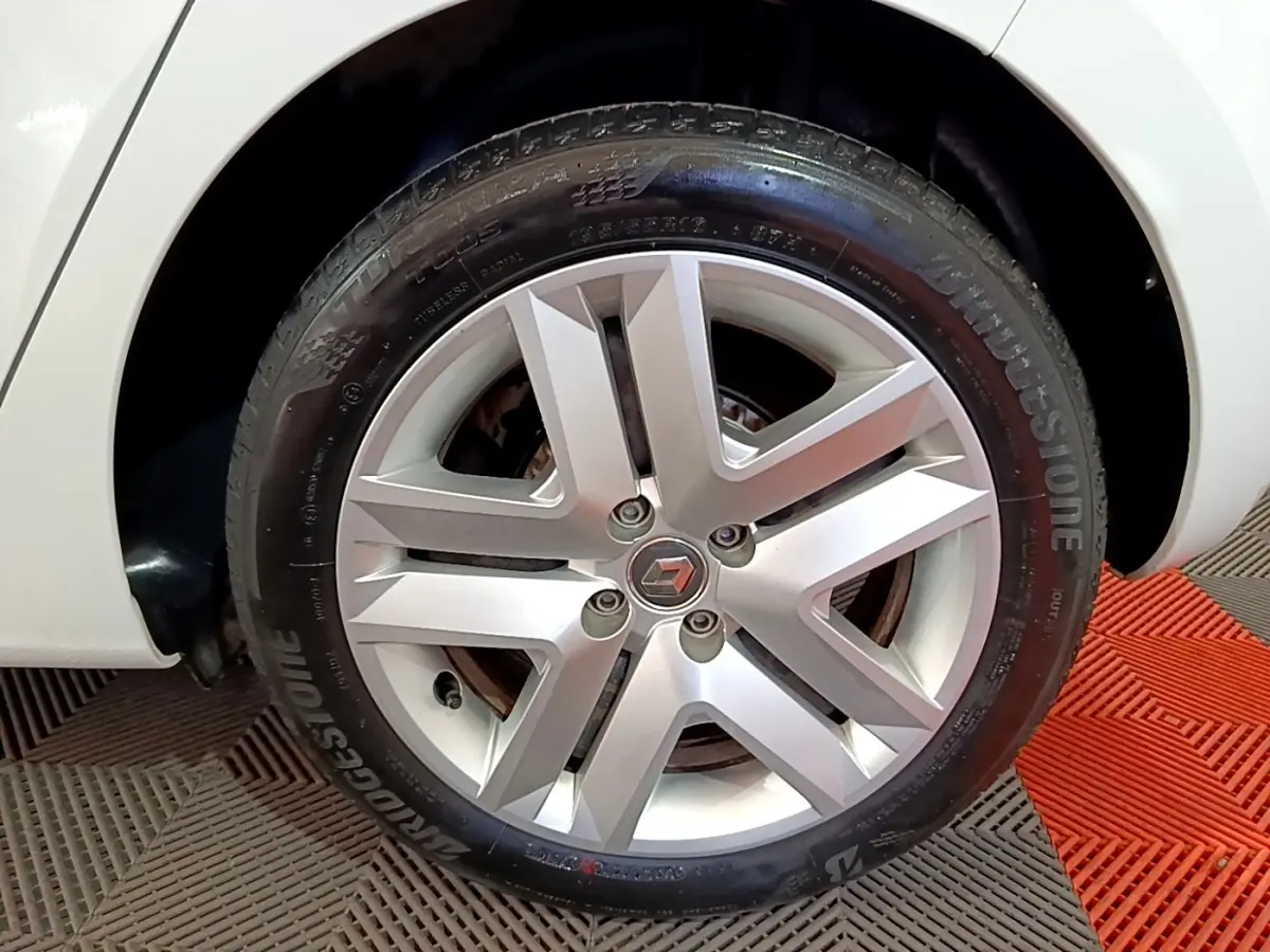 Gros plan sur la roue arrière droite d'une Renault Clio blanche, avec passage de roue visible et sol en damier rouge et gris.