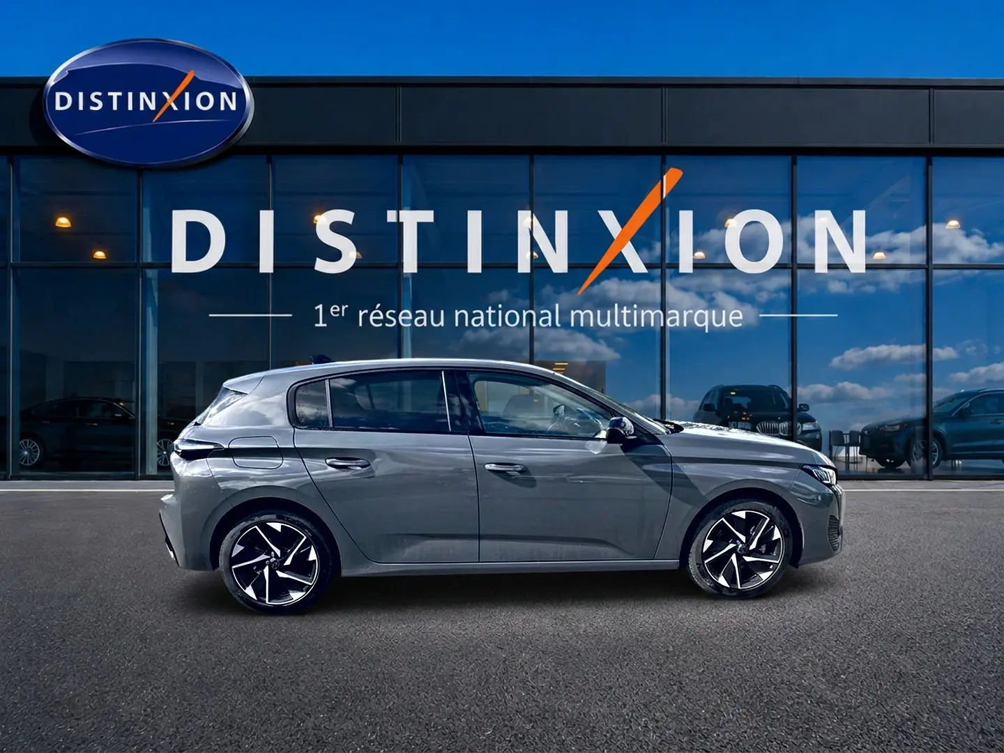 Profil droit d'une Peugeot 308 gris métallisé 2025 avec jantes alliage noires et blanches, devant un showroom Distinxion.