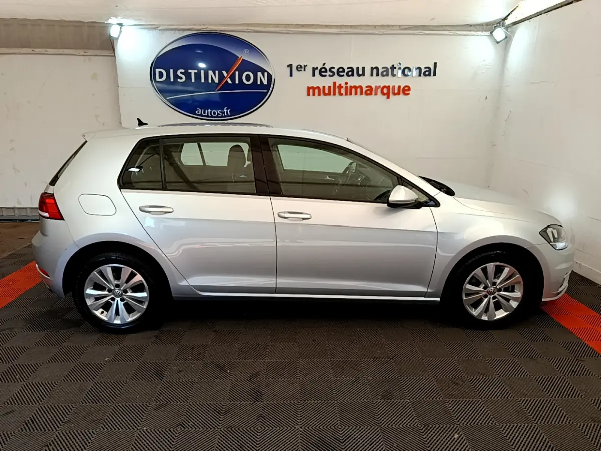 Profil côté gauche d'une Volkswagen Golf gris clair 2019 garée en intérieur avec logo Distinxion en arrière-plan.