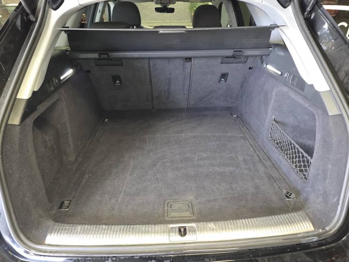 Coffre ouvert vu de l'arrière d'une Audi A4 Avant noire, avec tapis et filet de rangement latéral.