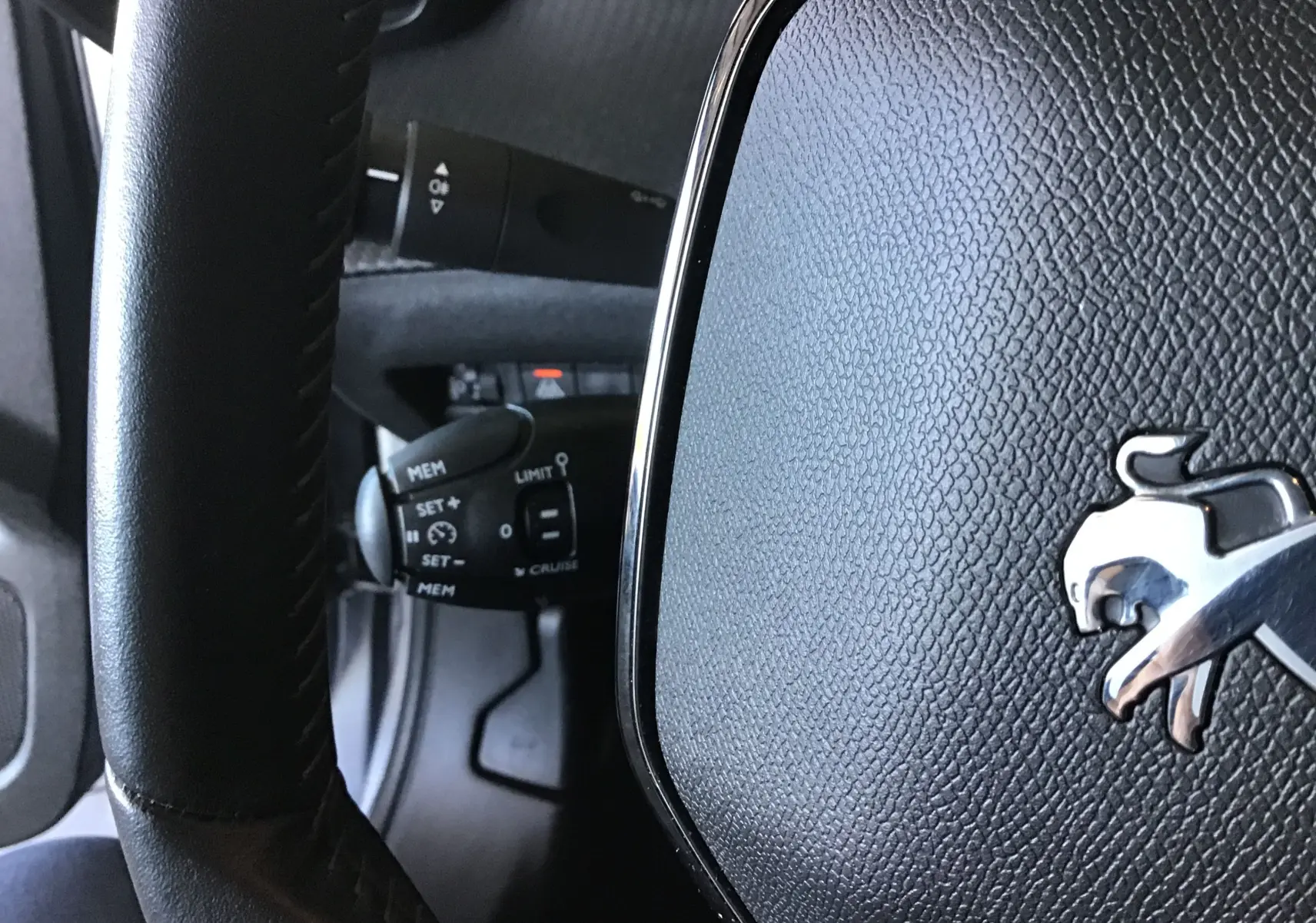 Gros plan sur le volant noir en cuir avec logo Peugeot argenté et commandes de régulateur de vitesse visibles derrière.