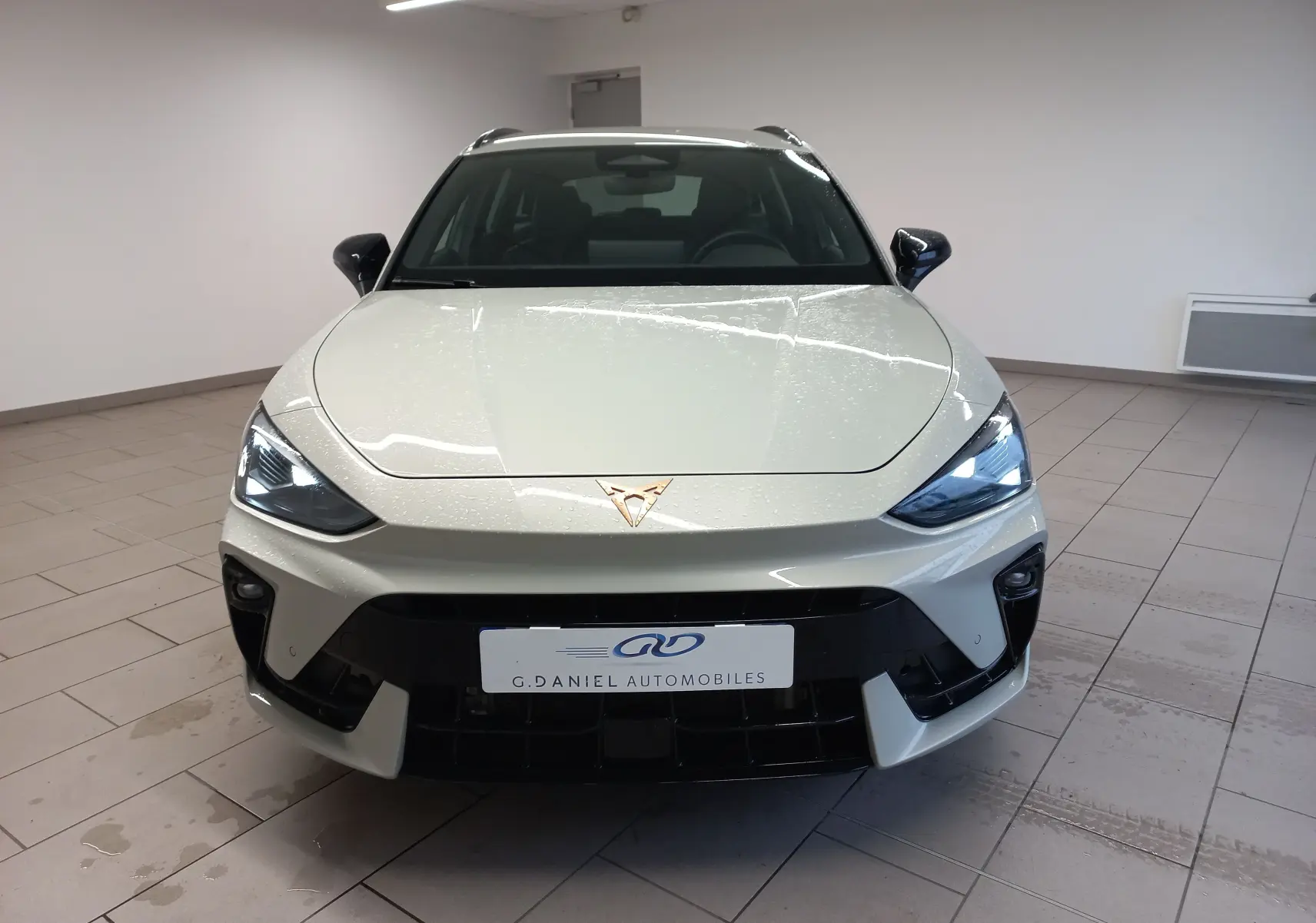 Vue avant d'une CUPRA LEON ST 1.5 eTSI 150 DSG7 en gris Taiga avec logo cuivré et phares allumés en intérieur.