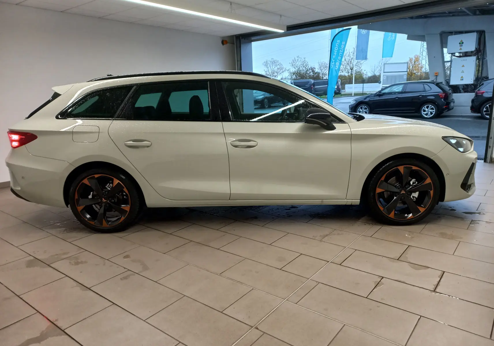Vue de profil droit d'une CUPRA LEON ST 1.5 eTSI 150 DSG7 en gris Taiga avec jantes alliage 18'' cuivrées dans un showroom.