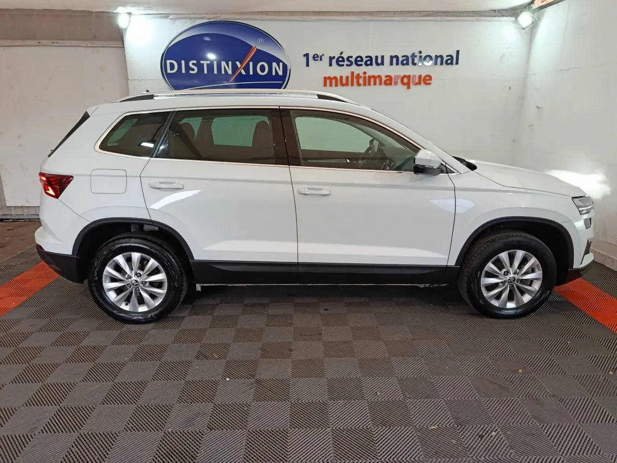 Vue latérale droite d'un SKODA KAROQ blanc 2024 garé en intérieur avec jantes alliage et bas de caisse noir.