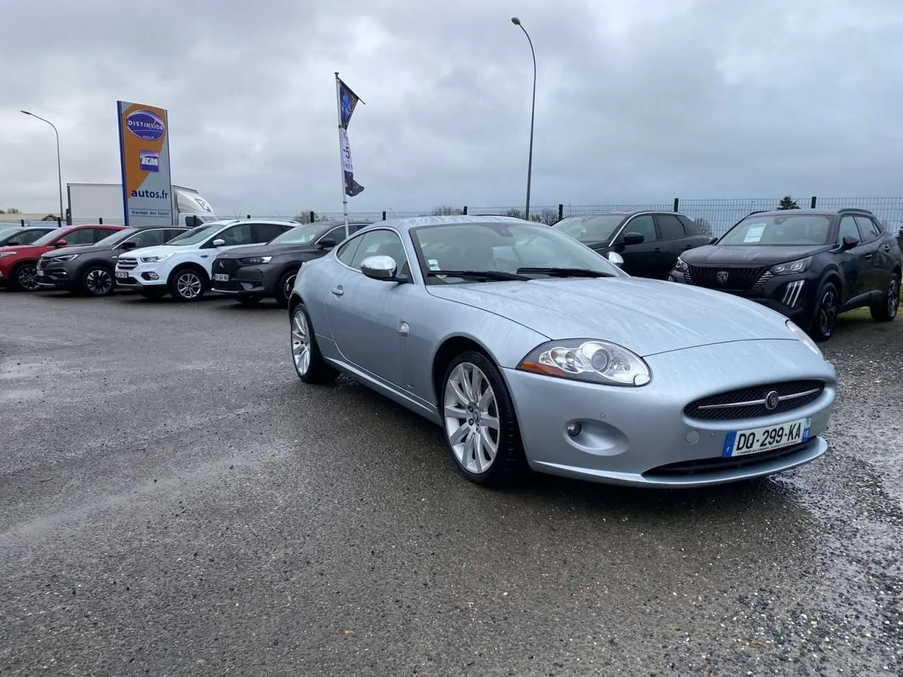 Jaguar XK Coupé gris clair vu en 3/4 avant droit, avec ses jantes alliage et calandre ovale distinctive.