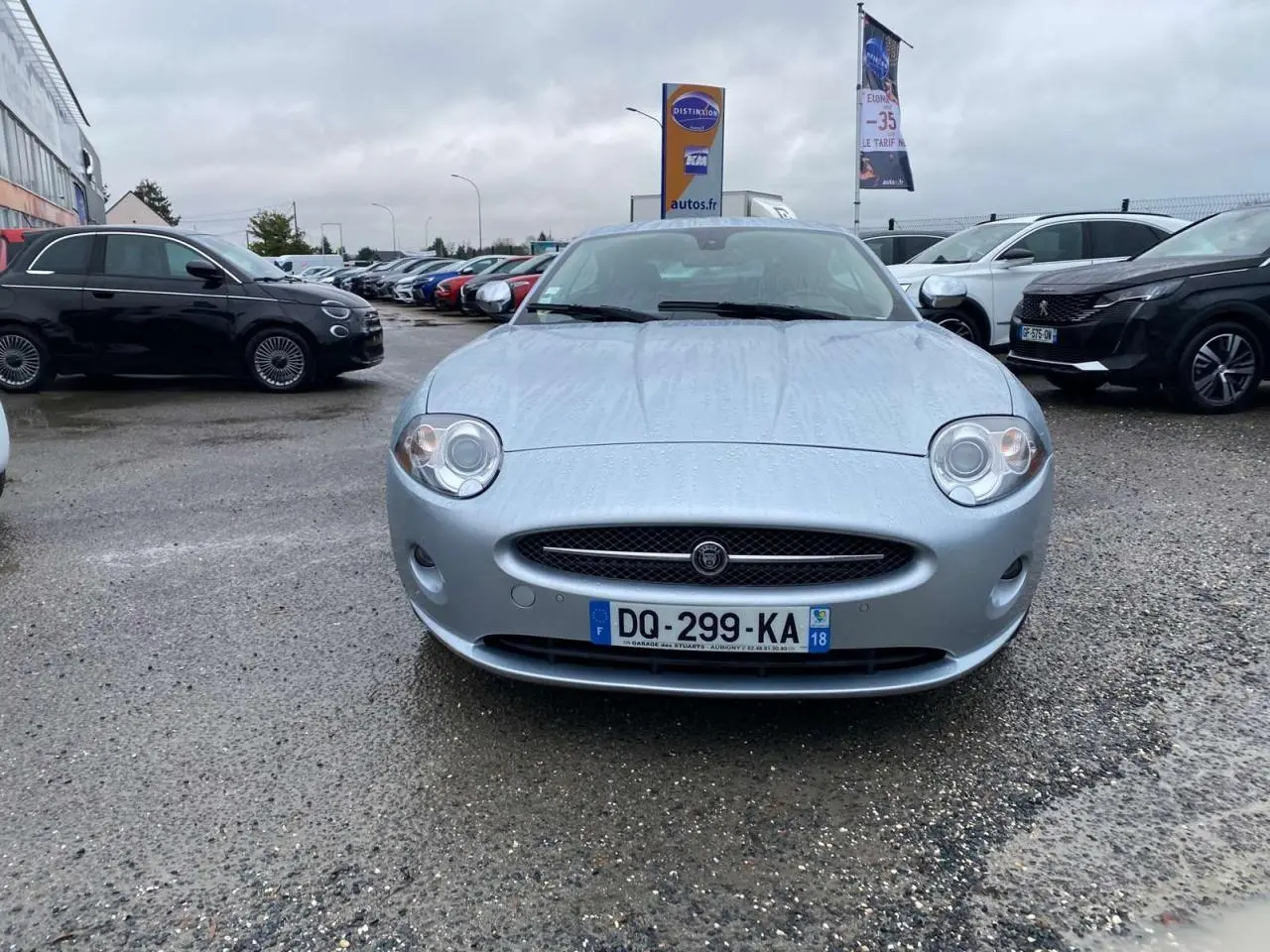 Jaguar XK Coupé gris clair vu de face sur un parking sous un ciel nuageux avec des gouttes de pluie visibles.