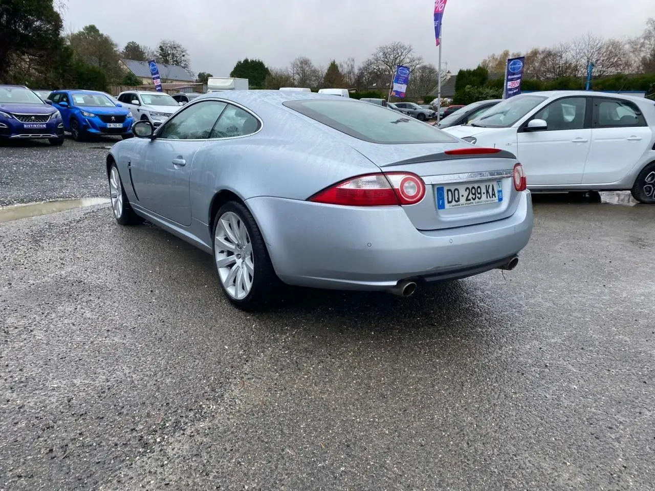 Jaguar XK Coupé gris clair vue 3/4 arrière droit, avec feux ronds caractéristiques et double sortie d'échappement.