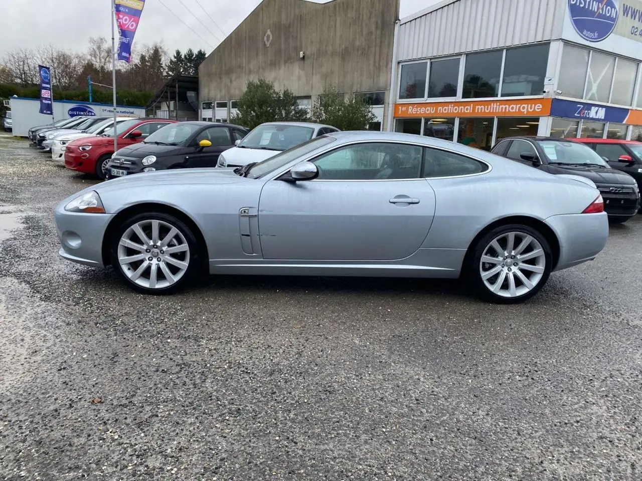 Jaguar XK Coupé gris clair vu de profil côté gauche, stationné devant un bâtiment commercial.