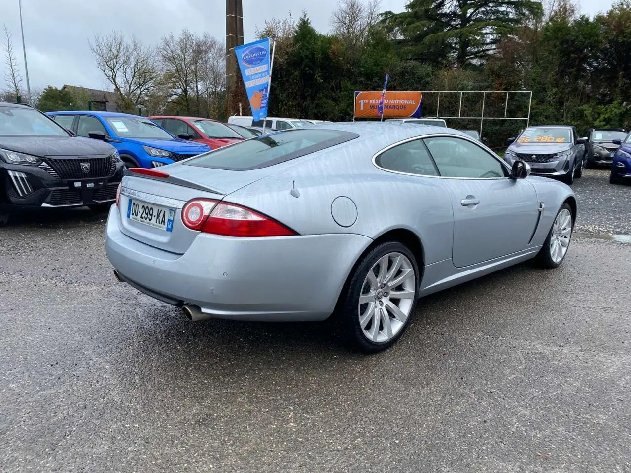 Jaguar XK Coupé gris clair vu 3/4 arrière droit, avec jantes alliage et feux arrière ronds distinctifs.