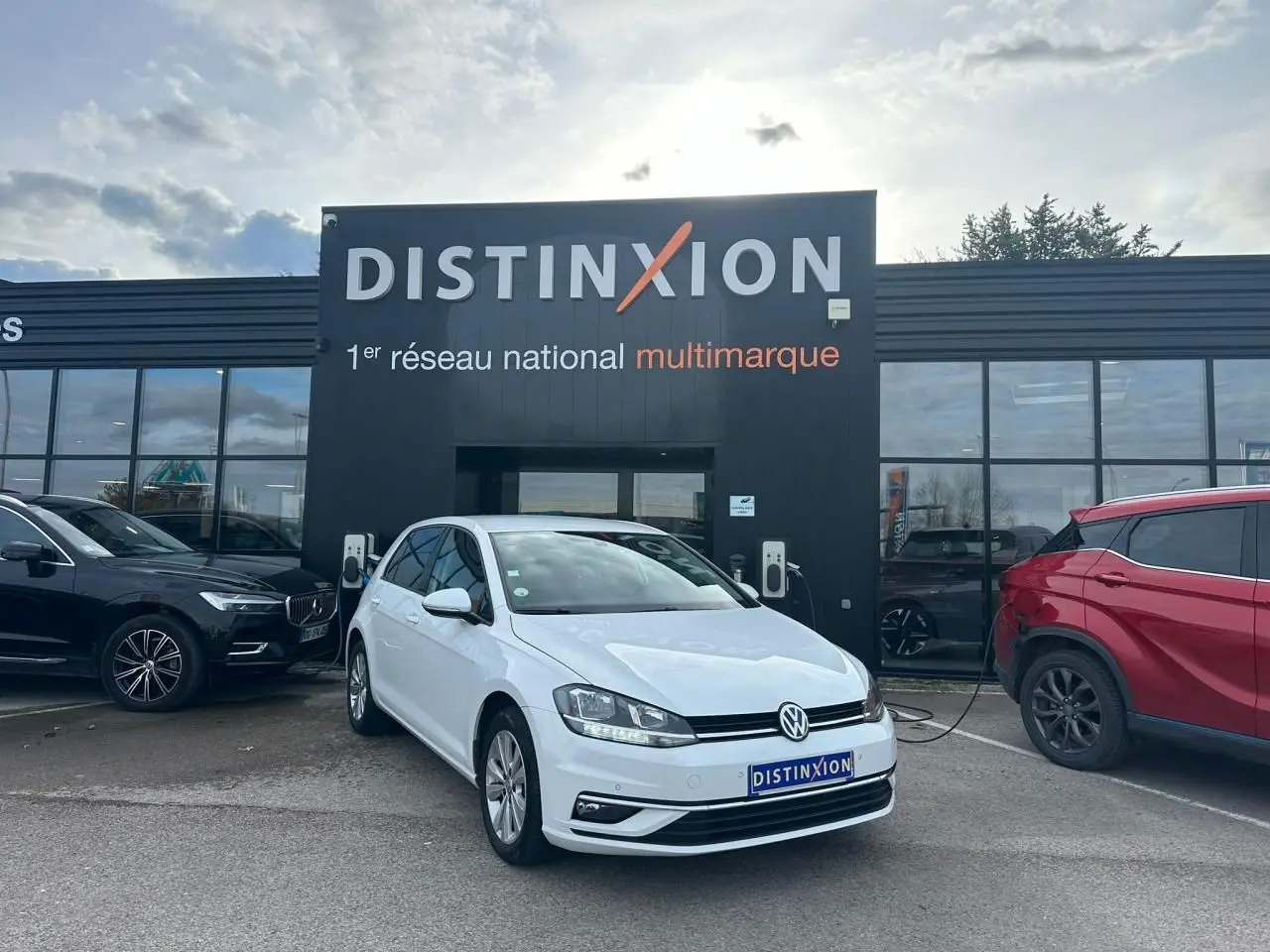Volkswagen Golf VII blanche vue 3/4 avant droit, stationnée devant un showroom Distinxion avec borne de recharge.