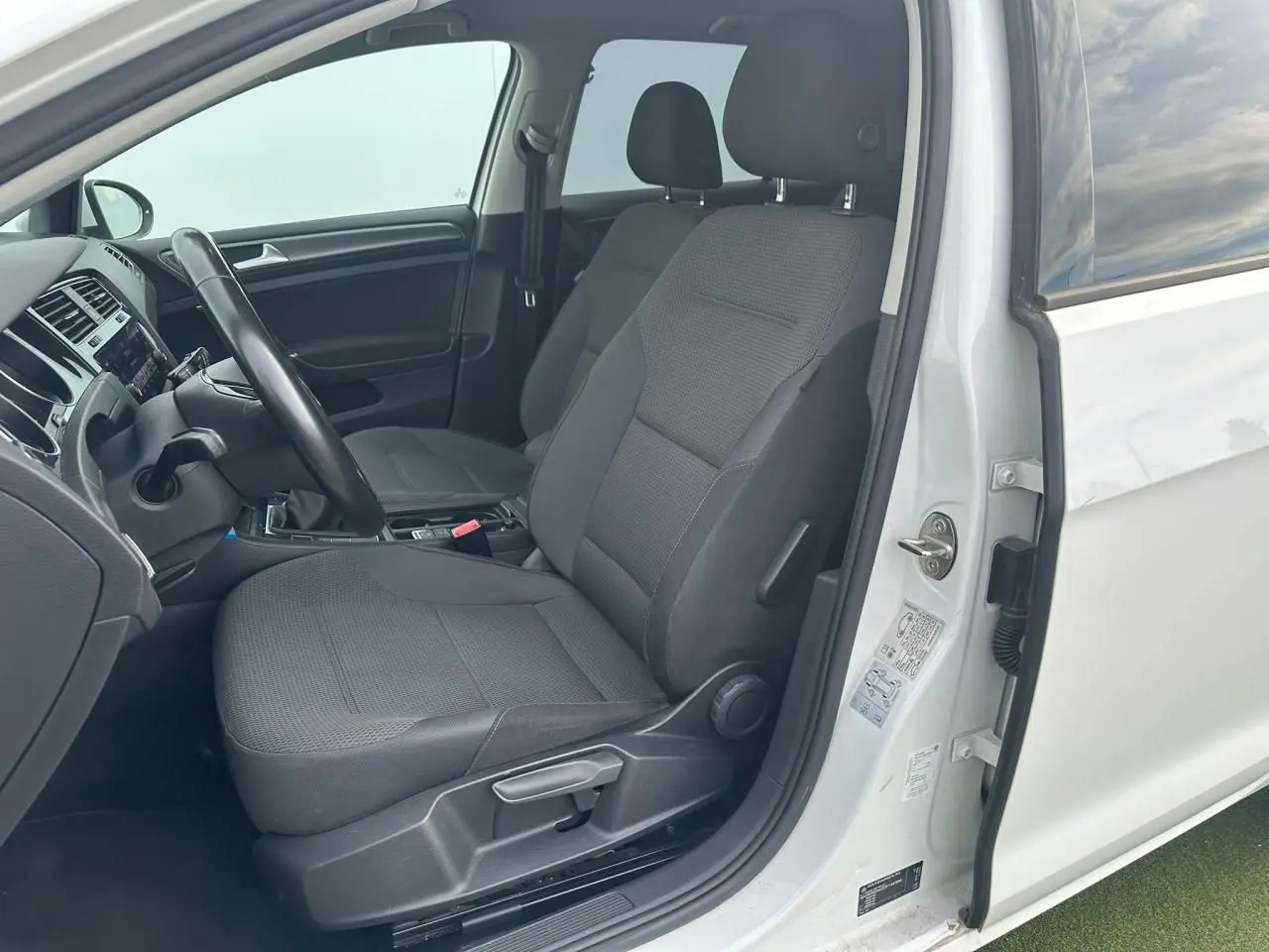 Vue intérieure côté conducteur de la Volkswagen Golf VII blanche, montrant les sièges avant en tissu gris et le volant.