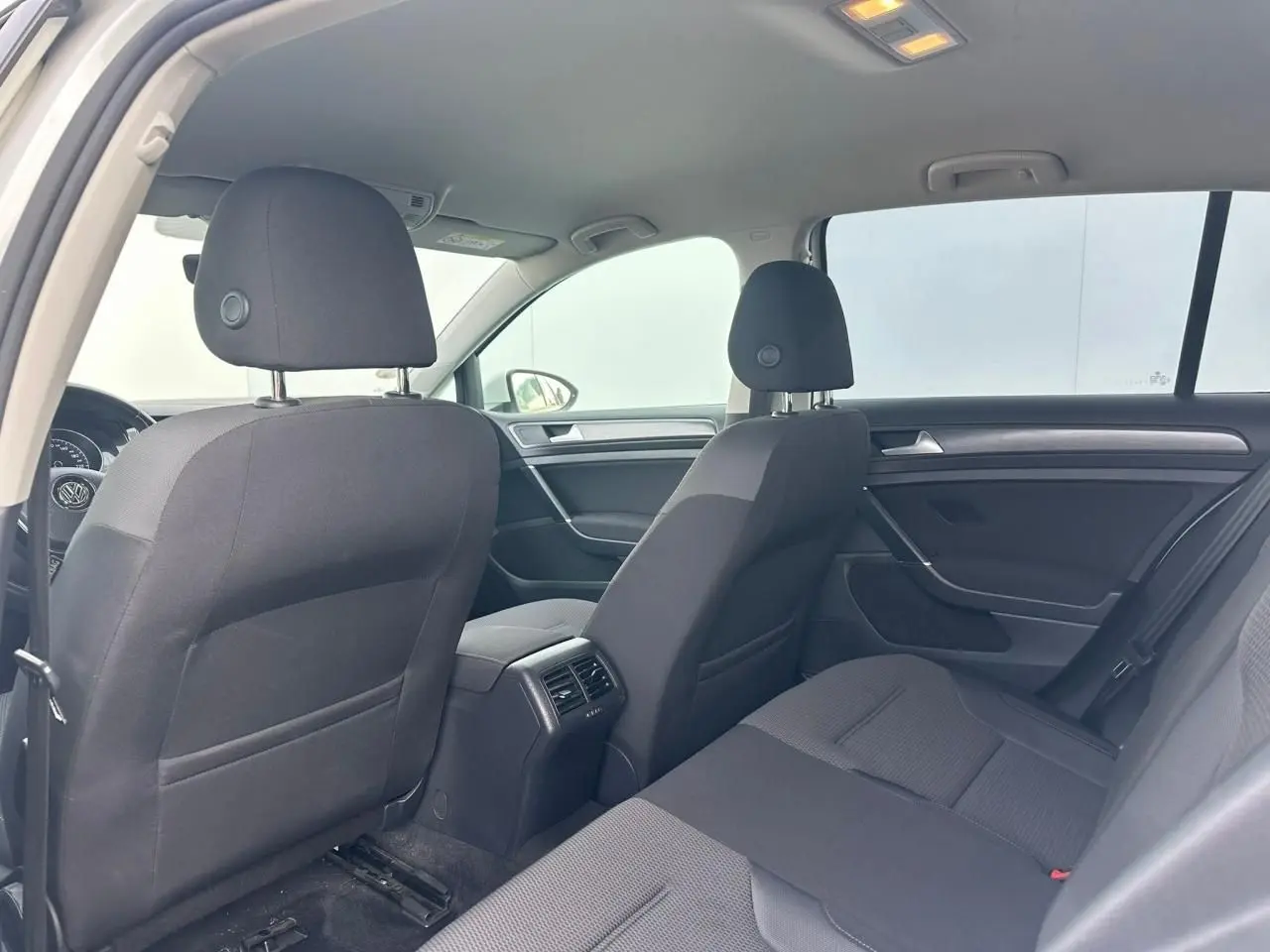 Vue intérieure arrière côté gauche de la Volkswagen Golf VII blanche, montrant les sièges en tissu noir et les aérateurs centraux.