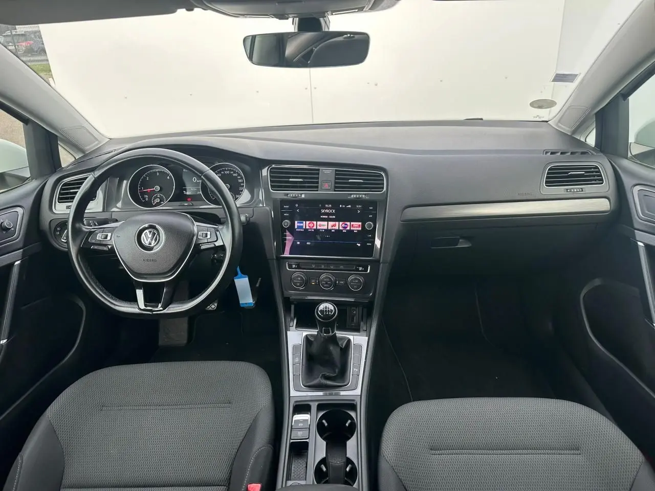 Vue intérieure avant de la Volkswagen Golf VII blanche, tableau de bord noir avec console centrale et volant cuir multifonction.