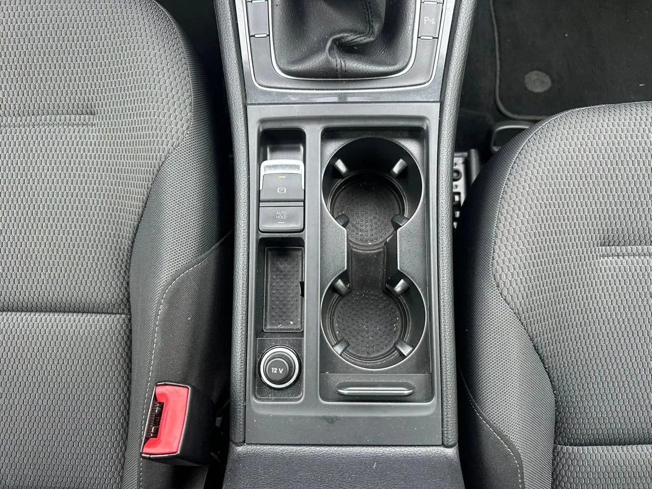 Vue plongeante sur la console centrale noire de la Volkswagen Golf VII blanche, avec porte-gobelets et levier de vitesses manuel.
