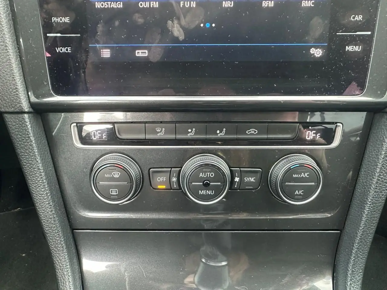 Gros plan sur la console centrale noire de la Volkswagen Golf VII blanche, affichant les commandes de climatisation automatique.