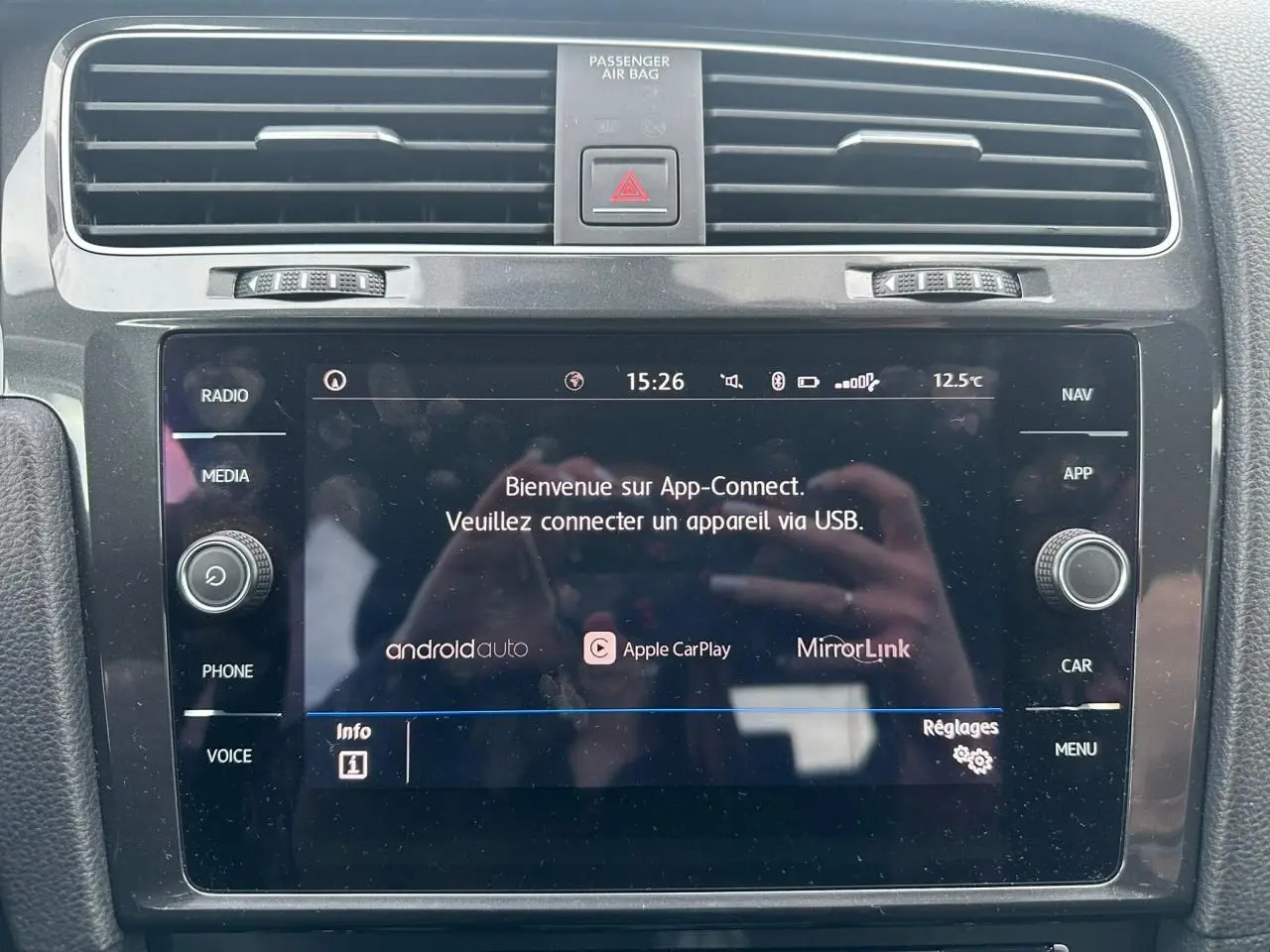 Gros plan sur l'écran tactile central noir de la Volkswagen Golf VII blanc, affichant App-Connect et commandes autour.