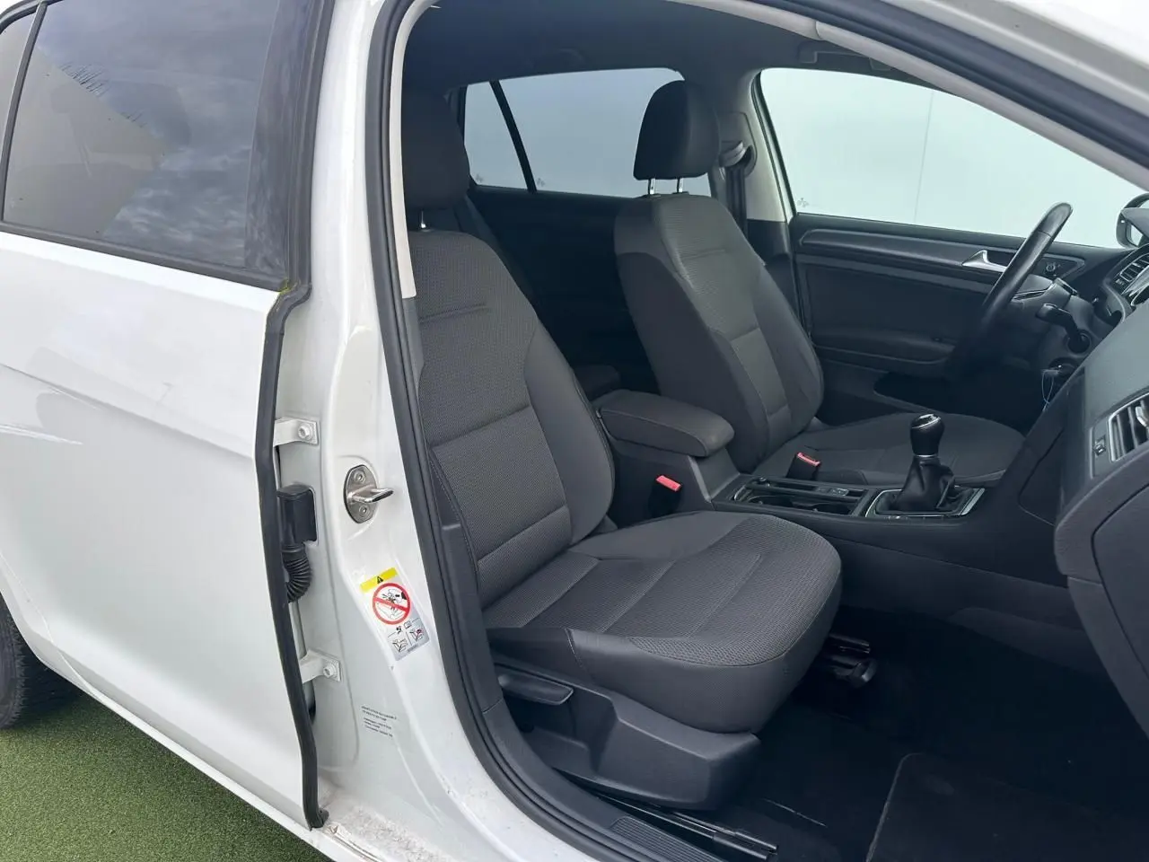 Vue intérieure côté gauche avant d'une Volkswagen Golf blanche avec sièges tissu gris et levier de vitesse manuel.