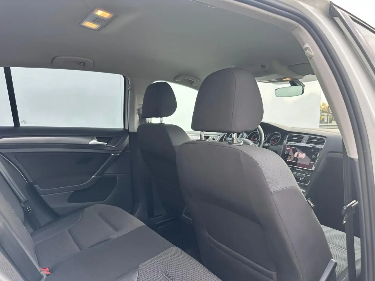 Vue intérieure arrière côté gauche de la Volkswagen Golf VII blanche, montrant les sièges en tissu noir et le tableau de bord avant.