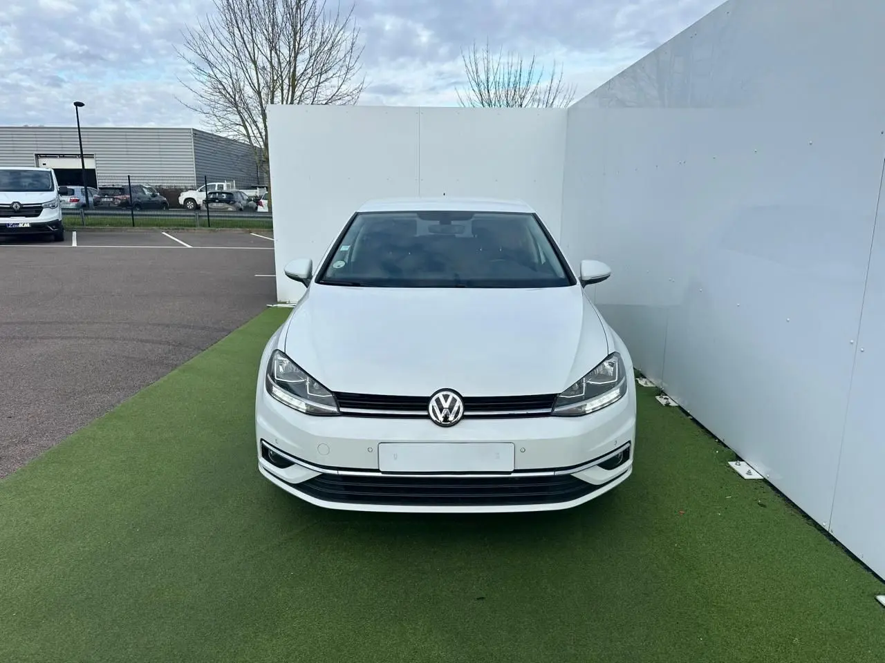 Vue frontale d'une Volkswagen Golf VII blanche garée sur un sol vert, avec calandre noire et logo VW visible.
