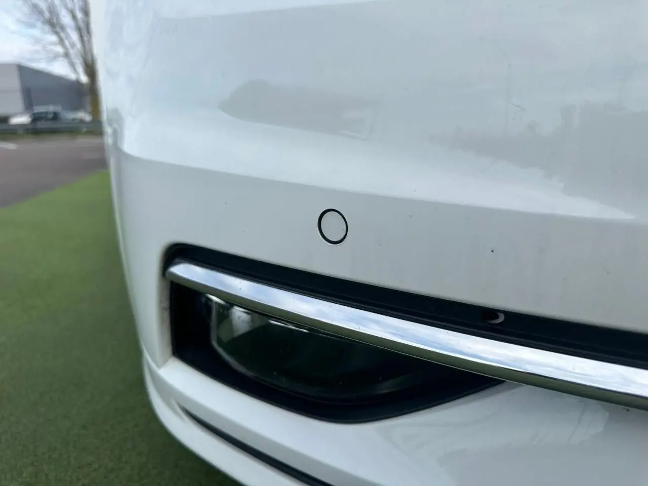 Gros plan sur le pare-chocs avant blanc d'une Volkswagen Golf VII avec capteur de stationnement et insert chromé.