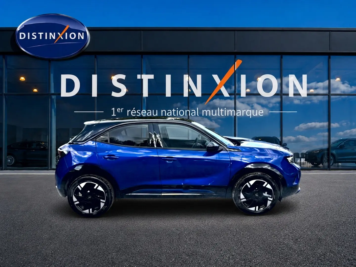 Opel Mokka Electric bleu en profil droit, avec jantes alu noires et toit noir devant une vitrine Distinxion.