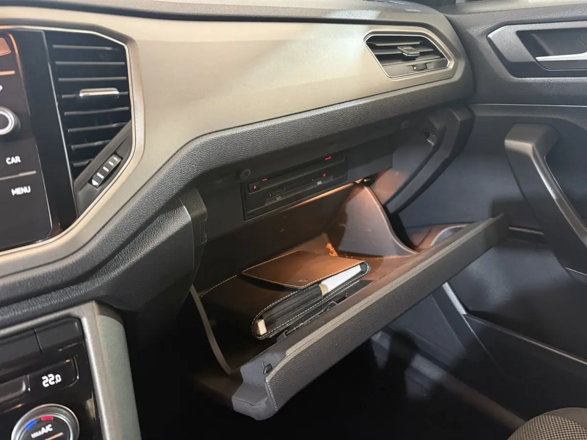 Vue rapprochée de la boîte à gants ouverte du Volkswagen T-Roc noir, mettant en valeur son intérieur et rangement éclairé.