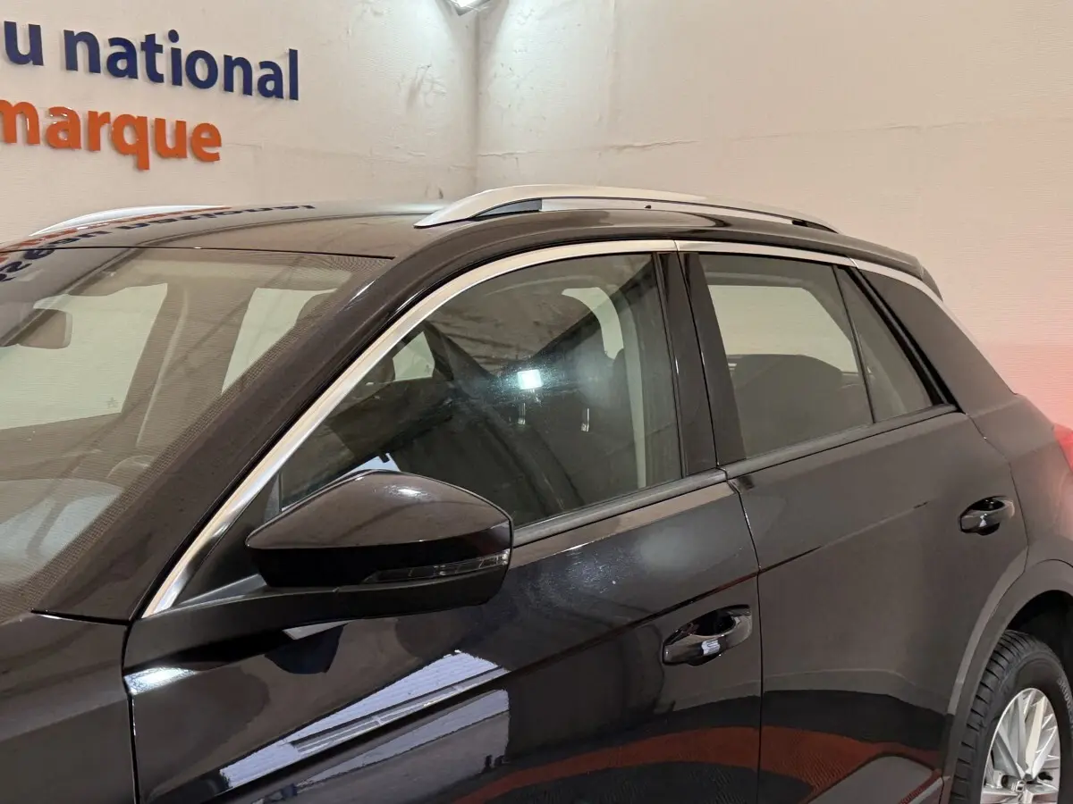 Vue latérale côté gauche d'un Volkswagen T-Roc noir 2020 avec barres de toit argentées en intérieur showroom.