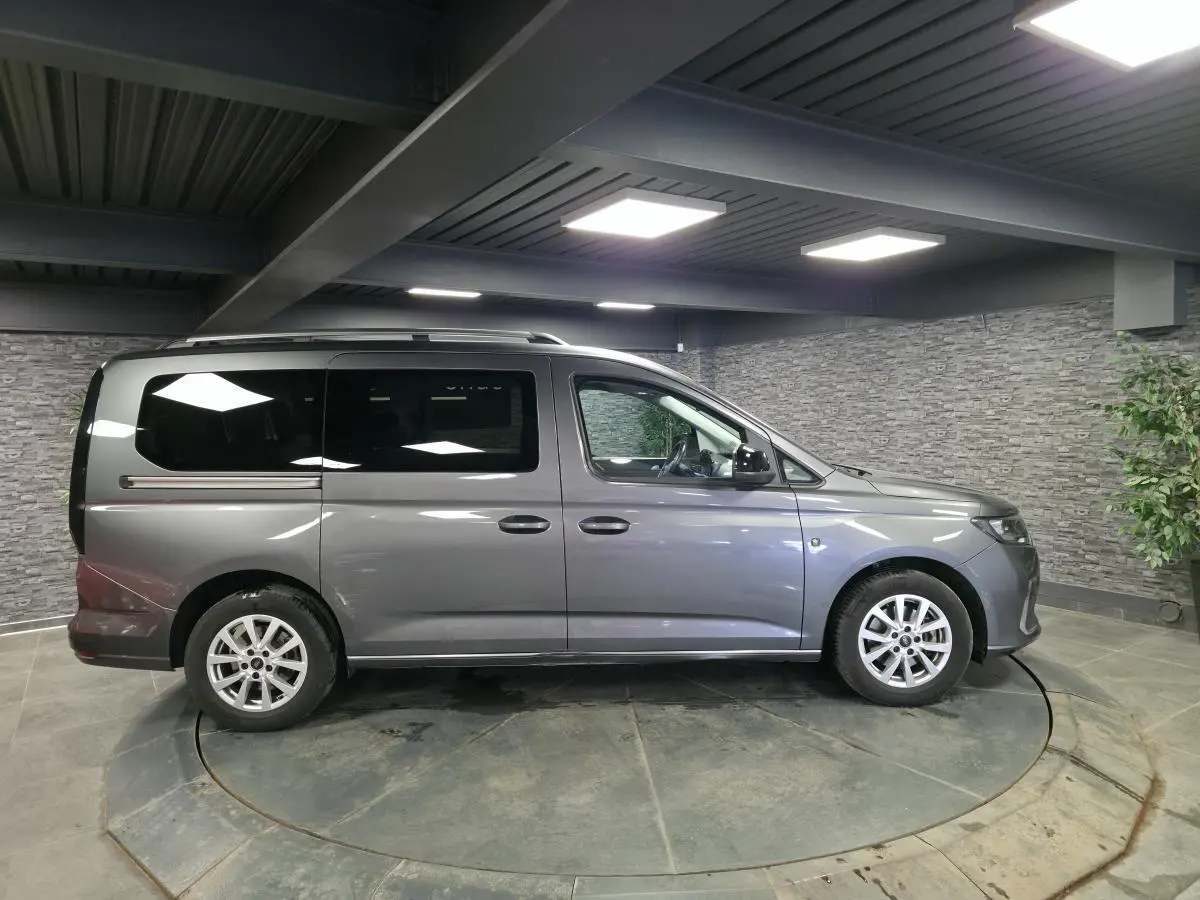 Vue latérale droite d'un Ford Transit Connect gris métallisé avec barres de toit argentées dans un showroom.