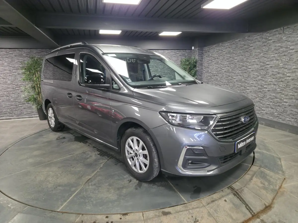 Vue 3/4 avant droit d'un Ford Transit Connect gris avec calandre noire et barres de toit argentées en intérieur showroom.