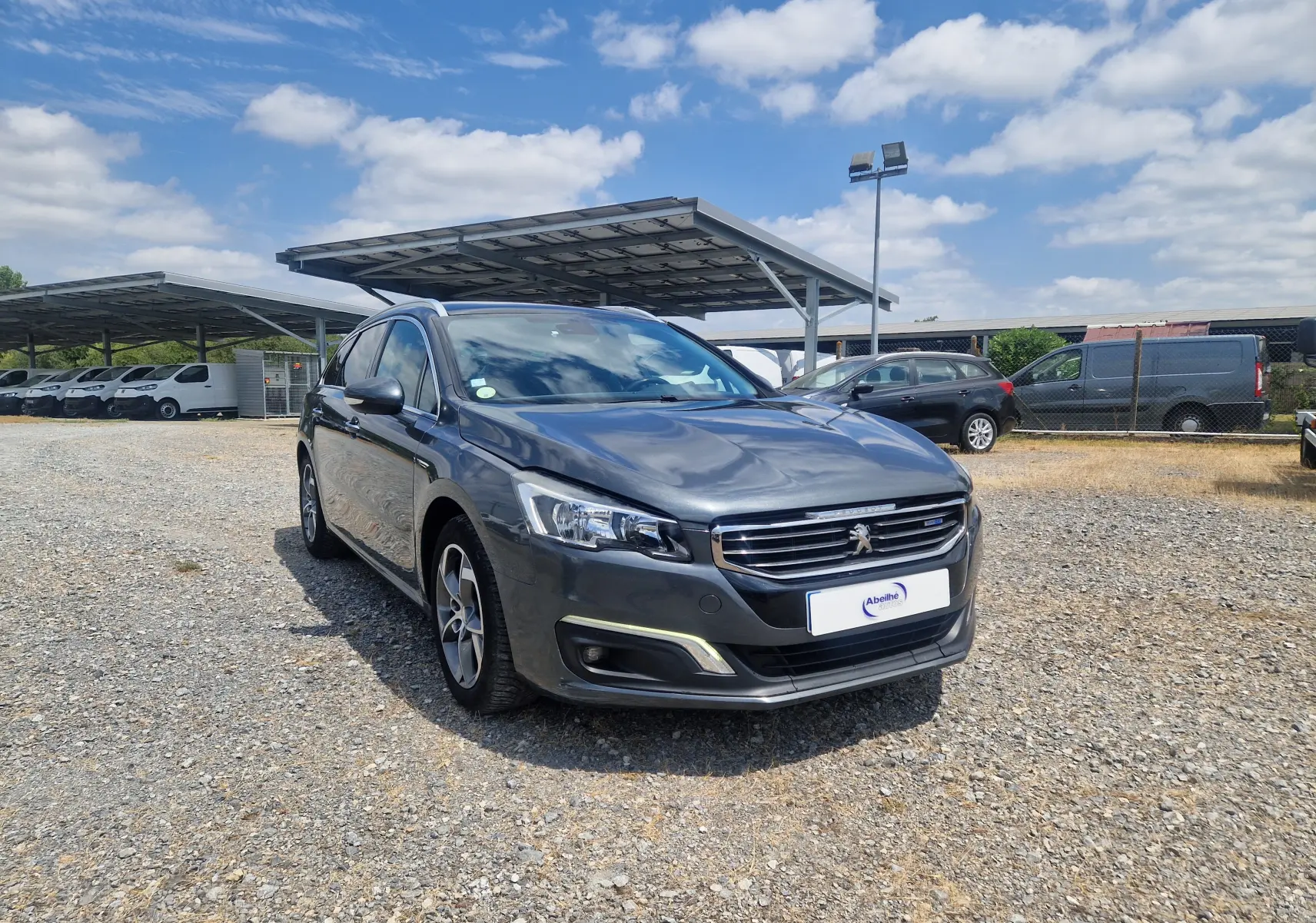 Peugeot 508 SW gris shark vue 3/4 avant droit sur un parking avec abris solaires en arrière-plan.