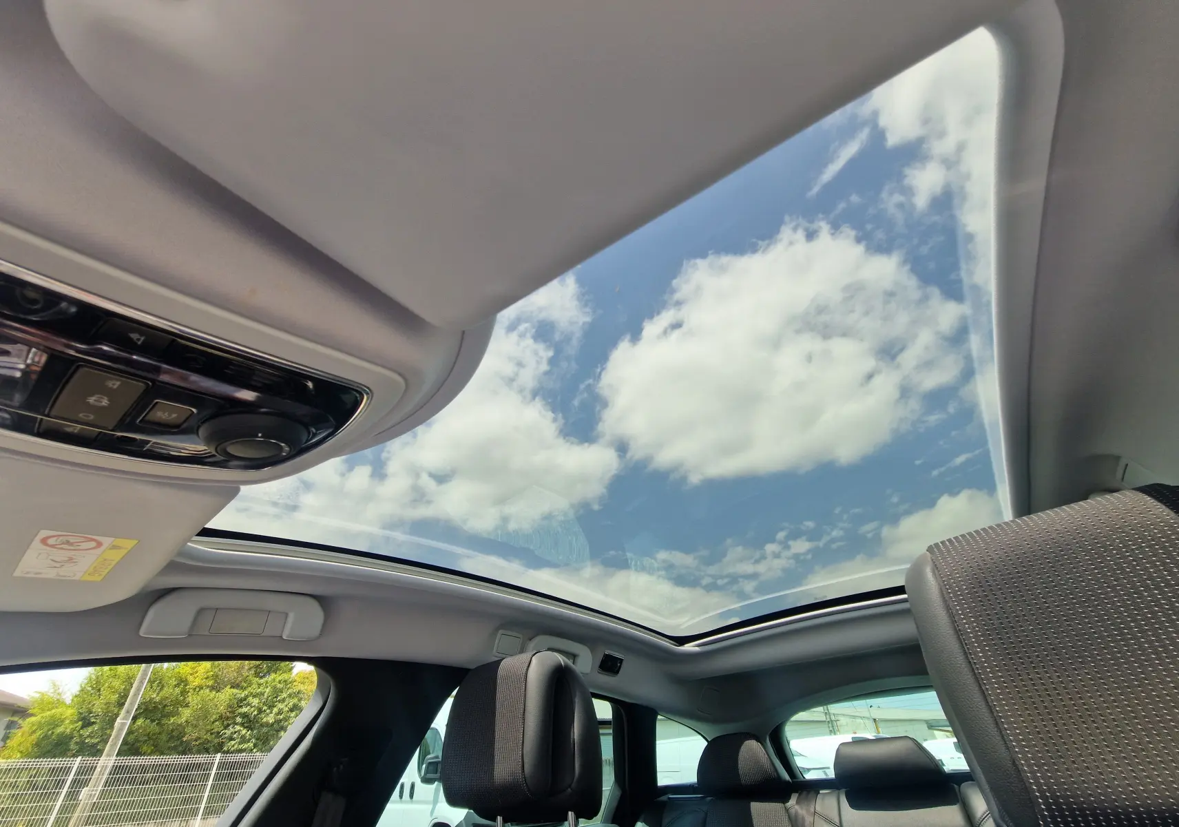 Vue intérieure du toit panoramique ouvrant du Peugeot 508 SW gris Shark, avec ciel bleu et nuages visibles.