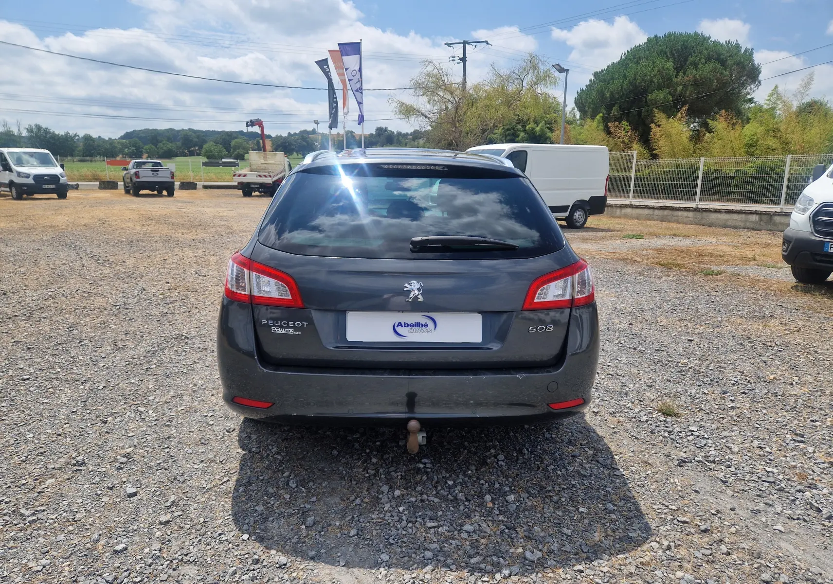 Vue arrière d'un Peugeot 508 SW gris shark de 2015 avec attelage, sur un terrain gravillonné en extérieur.