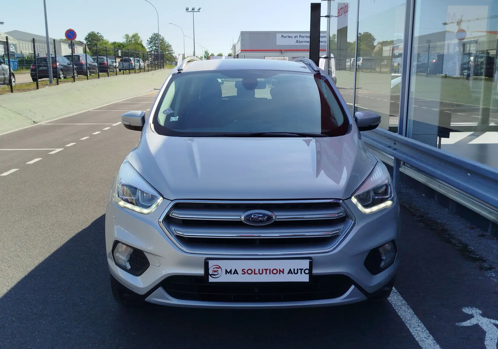 Vue frontale d'un Ford Kuga gris clair garé devant un bâtiment vitré avec phares allumés.