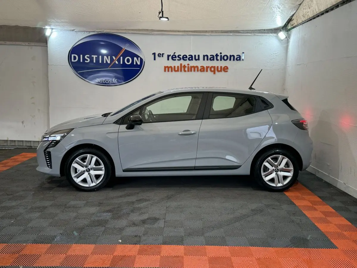 Profil côté gauche d'une Renault Clio gris clair 2025 dans un showroom avec éclairage intérieur et logo Distinxion en arrière-plan