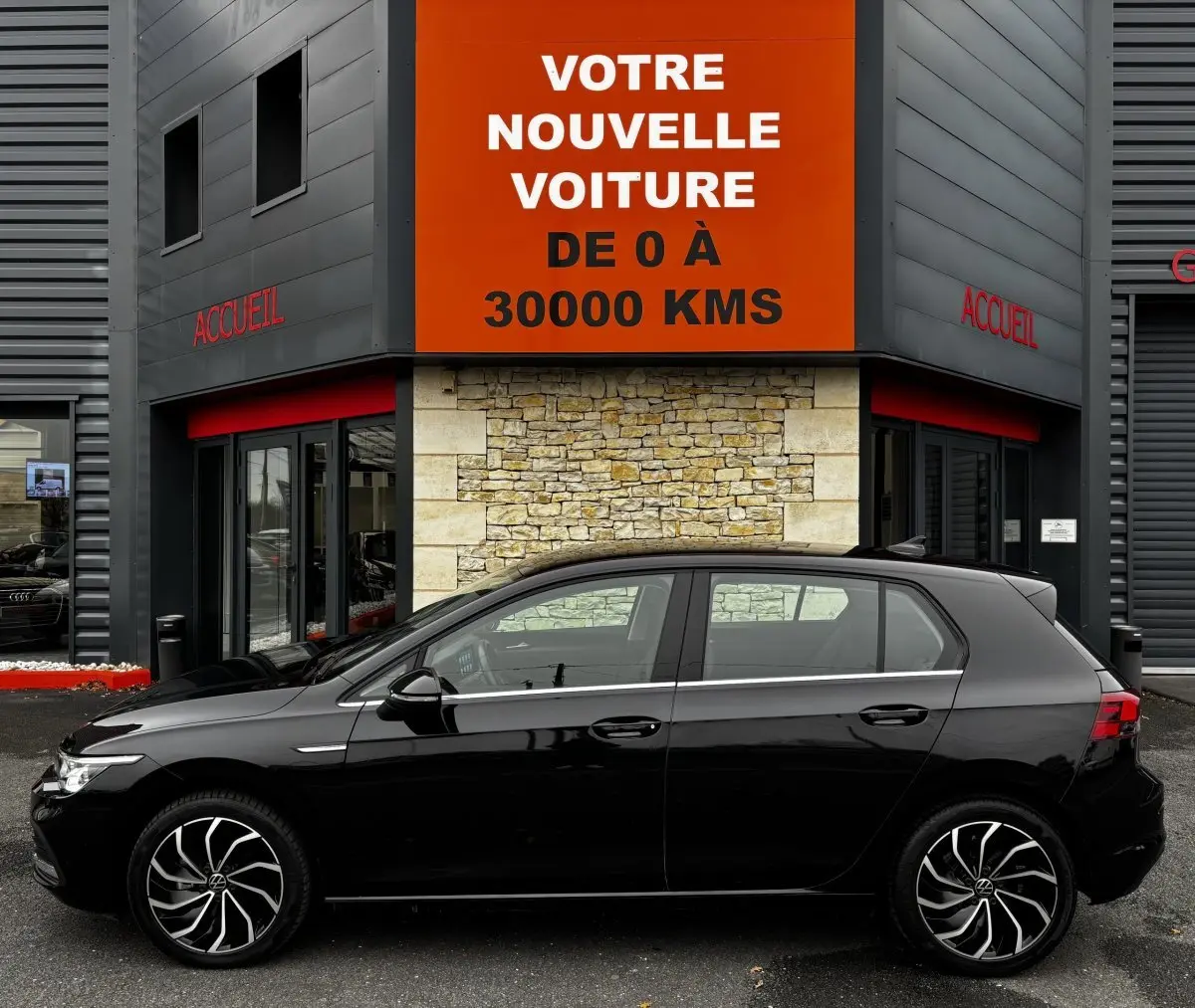 Profil côté gauche d'une Volkswagen Golf noire 2024 garée devant un bâtiment avec panneau orange "Votre nouvelle voiture".