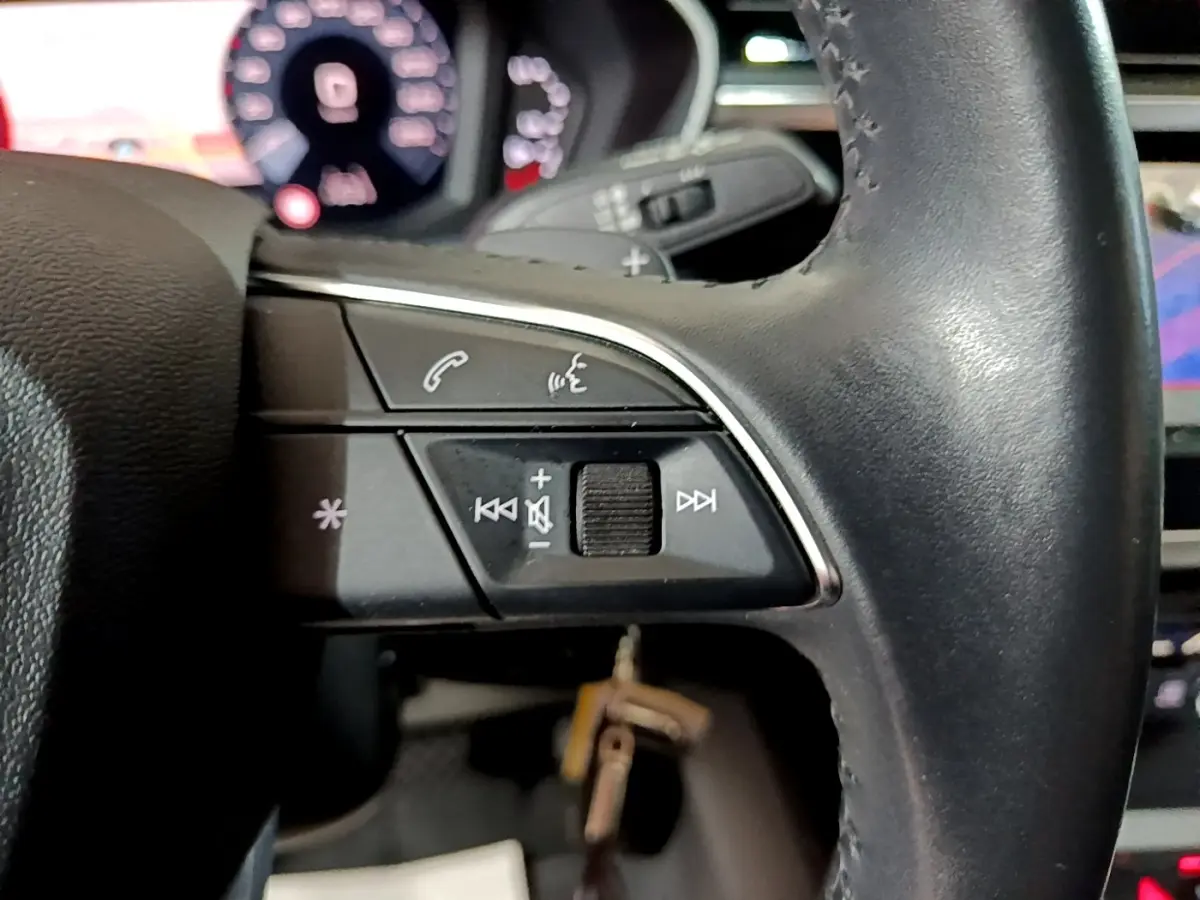 Gros plan sur les commandes du volant cuir noir de l'Audi Q3 35 TDI 150 S tronic Business Line, avec tableau de bord numérique flou en arrière-plan.